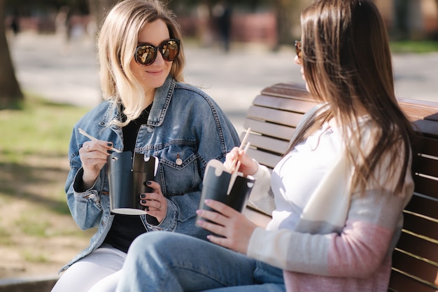 Amigas comem comida asiática para viagem, sentadas em um banco