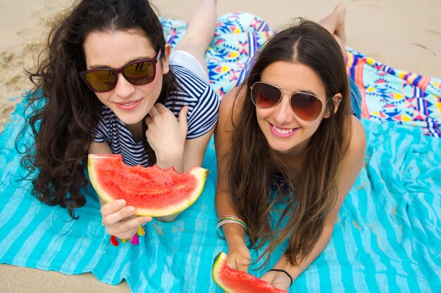 Amigas comendo melancia enquanto estão deitadas em um cobertor na praia