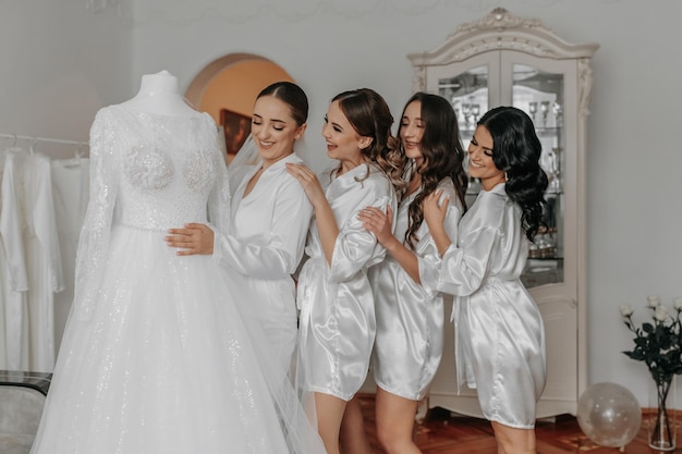 Foto amigas da noiva junto com a noiva perto do vestido de noiva a noiva e seus amigos divertidos celebram a despedida de solteira em vestidos correspondentes noiva e amigos no quarto