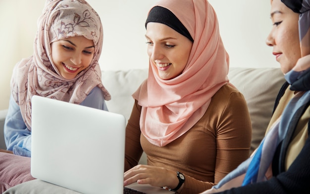 Amigas islámicas sentadas en el sofá hablando y usando una computadora portátil