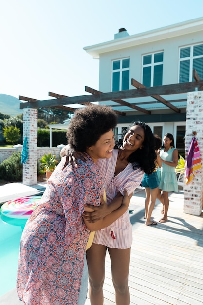 Amigas milenarias birraciales despreocupadas abrazándose y disfrutando de las vacaciones de verano junto a la piscina