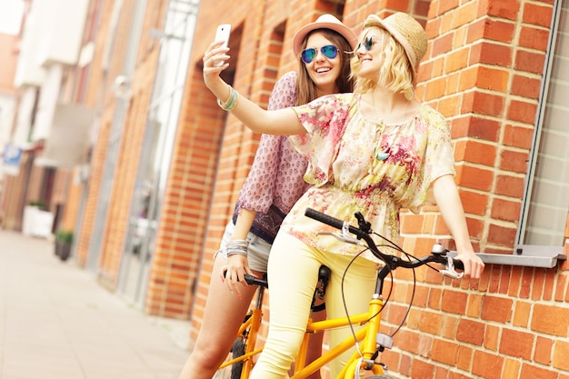 amigas montando una bicicleta tándem y tomando selfie en la ciudad