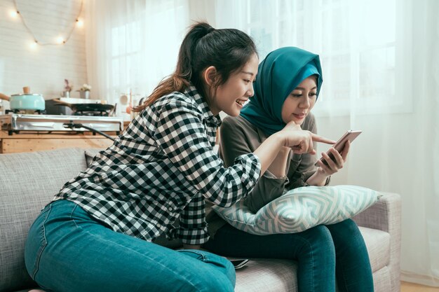 Amigas multiétnicas eufóricas assistindo vídeo no smartphone e apontando a tela surpresa. sorridente garota japonesa sentada com o colega de quarto do Islã no sofá no apartamento moderno e brilhante usando o celular