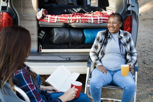 Amigas multirraciales que se divierten acampando con autocaravana mientras leen y toman café al aire libre Enfoque en la cara mayor africana