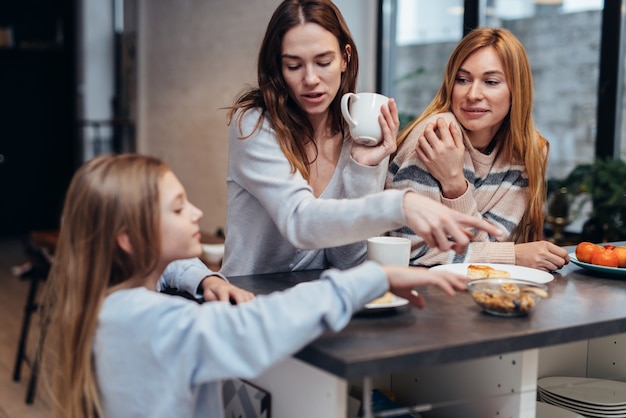 Amigas y una niña sentados juntos en la mesa de la cocina, bebiendo té y hablando.