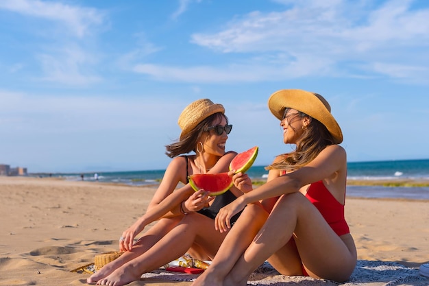 Amigas de vacaciones en la playa en verano comiendo una sandía con el mar de fondo sentadas en la arena