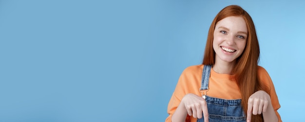 Amigável feliz garota ruiva sorrindo animado sorriso bonito apontando para baixo dedos indicadores oferecendo bom