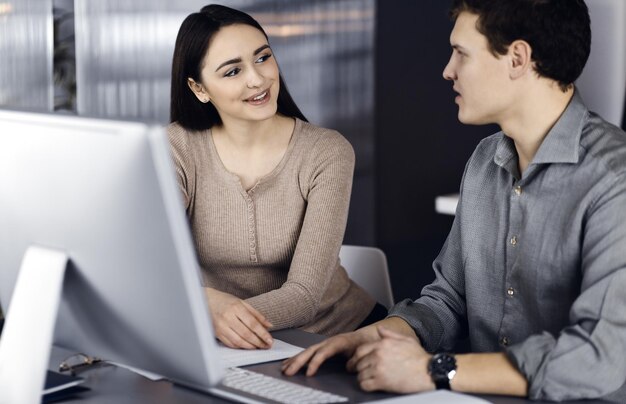 Amigável jovem empresário e programador de camisa verde está trabalhando no computador, enquanto está sentado junto à mesa com uma colega em um escritório moderno. Foco na mulher. Conceito de sucesso