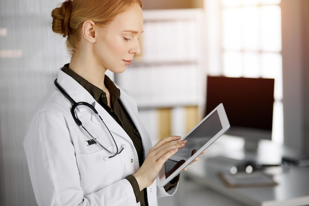 Amigável médica sorridente usando computador tablet na clínica ensolarada mulher médica no trabalho