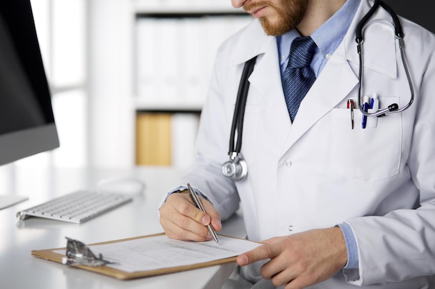 Amigável médico barbudo ruivo sentado e escrevendo com prancheta na clínica, close-up conceito de medicina.