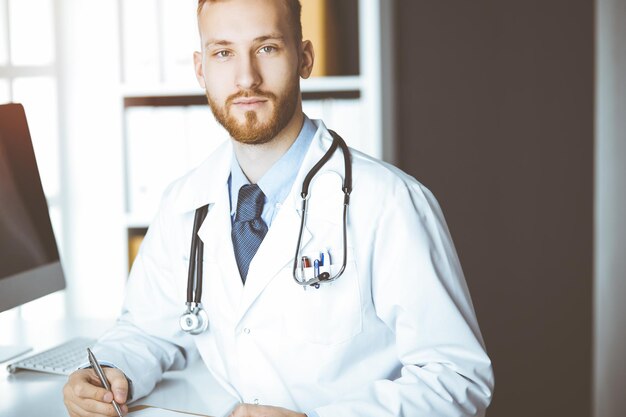 Amigável médico barbudo sentado e escrevendo na área de transferência na clínica ensolarada. conceito de medicina, parada pandêmica.