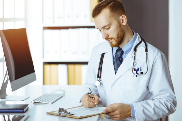 Amigável médico ruivo sentado e escrevendo na área de transferência na clínica ensolarada. Conceito de medicina.