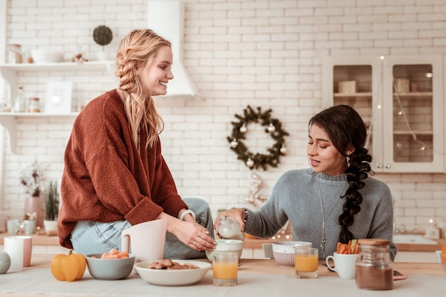 Amigo compartilhando leite. Namoradas atraentes tendo uma conversa agradável em uma cozinha leve e elegante enquanto saboreiam o café da manhã comum