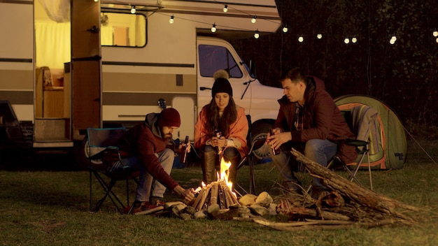 Amigo de una pareja que ayuda a encender la fogata. Furgoneta autocaravana retro. Bombillas de fondo.