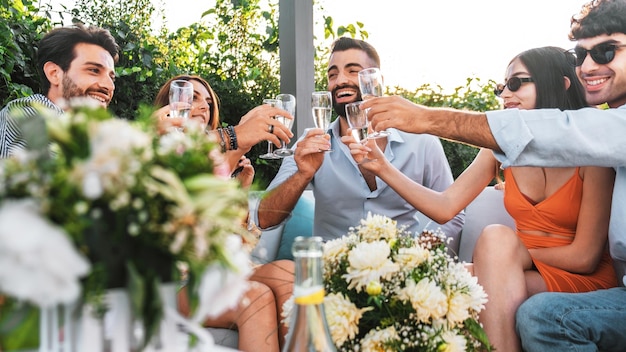 Amigo sparty reuniéndose en la terraza y brindando con champán sentados en el sofá - los mejores amigos se divierten bebiendo vino espumoso en el jardín de lujo - se centran en las copas