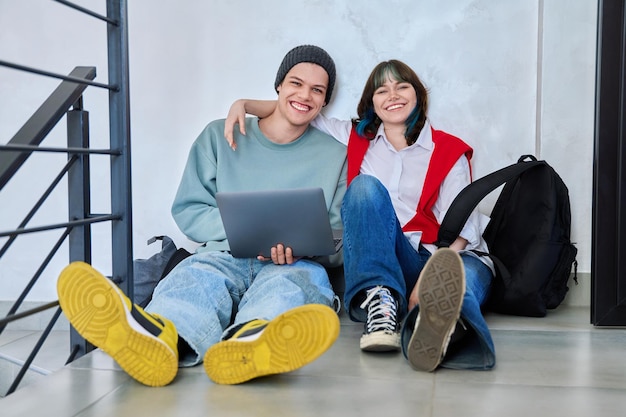 Amigos adolescentes hipster chico y chica sentados en el suelo con mochilas usando portátil