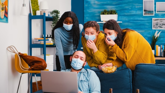 Amigos alegres de raza mixta mirando en una computadora portátil en un clip divertido sentado en la sala de estar con máscaras protectoras que mantienen el distanciamiento social para evitar la propagación del coronavirus