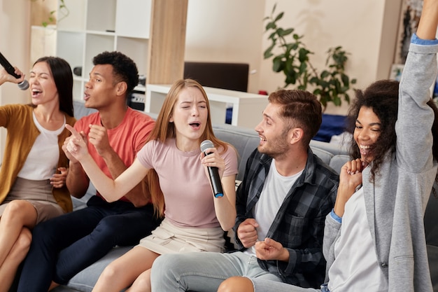 Amigos y amigos alegres de la música que parecen emocionados mientras juegan karaoke en casa cantando con