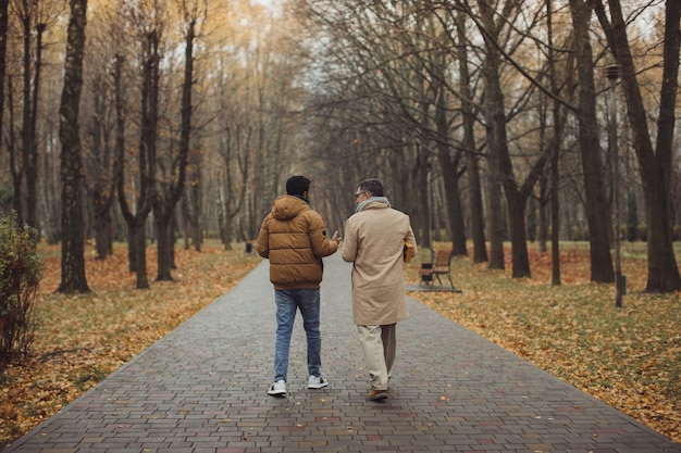 Amigos, un anciano y un joven caminando, hablando y tomando café juntos en el parque de otoñoxA