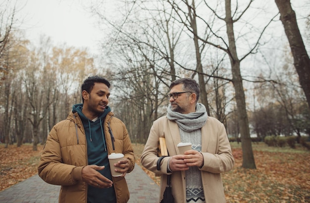 Amigos, un anciano y un joven caminando, hablando y tomando café juntos en el parque de otoñoxA