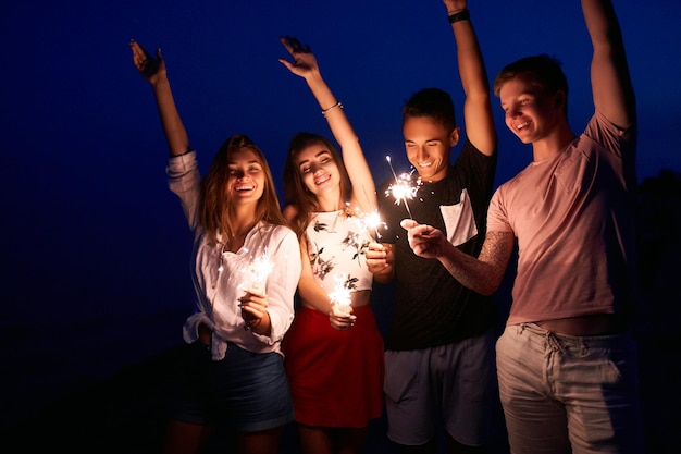 Amigos andando dançando e se divertindo durante a festa noturna à beira-mar com luzes de bengala nas mãos Jovens adolescentes festejando na praia com fogos de artifício Câmera lenta steadycam tiro