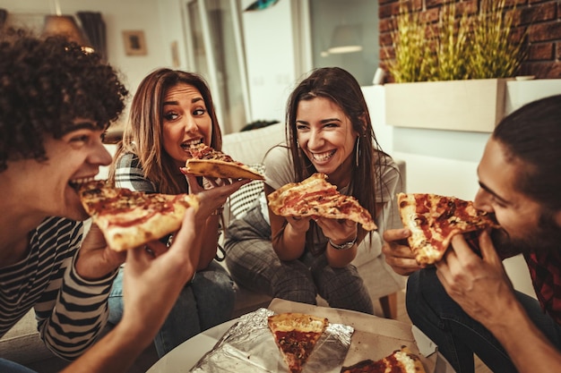 Amigos animados está em casa sentado no sofá. Seus amigos trazem pizza para comerem juntos. Eles têm um ótimo fim de semana em boa companhia dentro de casa.