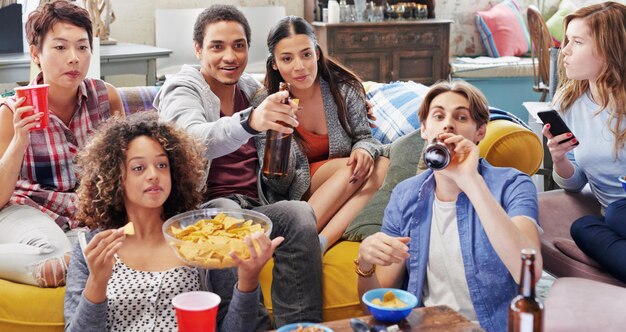 Los amigos beben y comen bocadillos y miran televisión, se relajan y cerveza en la sala de estar, diversidad y deportes juntos en casa. Hombres, mujeres y personas ven deportes de televisión y se suscriben a la transmisión con alcohol.