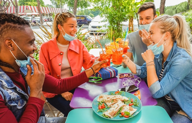 Amigos bebendo coktail e comendo petiscos de tapas em um bar de restaurante ao ar livre nos dias de verão com máscara facial para serem protegidos do coronavírus - pessoas felizes torcendo com spritz e se divertindo