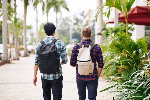 Amigos caminando por la calle con mochilas al explorar la nueva ciudad, vista desde atrás