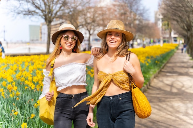 Amigos en la ciudad con sombreros de paja caminando y sonriendo junto a unas hermosas flores amarillas disfrutando de la primavera de vacaciones