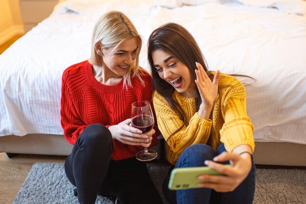 Amigos com vinho no chão em casa Amigos de mulheres jovens sorridentes felizes olhando fotos no celular Meninas se divertindo Duas amigas relaxando em casa com um copo de vinho conversando