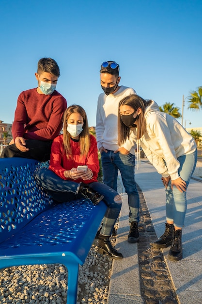 Amigos da geração Y usando smartphones usando máscara protetora na onda de Covid. Rapazes e raparigas preocupados a ver notícias no smartphone móvel. Grupo de viajantes mantém contato durante a pandemia nas férias