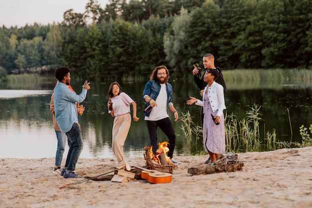 Amigos de estudantes se encontram na praia à beira do lago acendem uma fogueira fritar salsichas de pão tocam violão