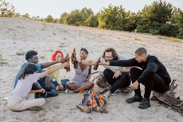 Amigos de longa data se encontram no lago na praia perto do fogo sentam na areia em toras de madeira bebem cerveja