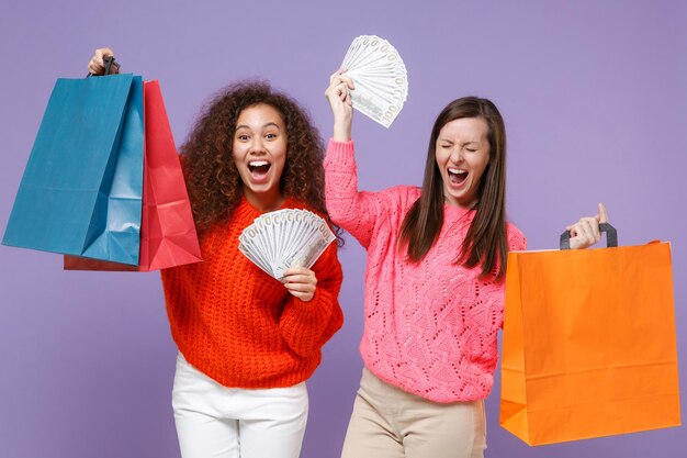 Foto amigos de mulheres afro-americanas europeias muito felizes em camisolas de malha isoladas em fundo roxo violeta. conceito de estilo de vida de pessoas. segure o saco do pacote com as compras após as compras, fã de dinheiro em espécie.