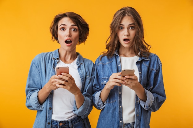 Amigos de mulheres jovens emocionais chocados posando isolados sobre uma parede amarela usando telefones celulares.