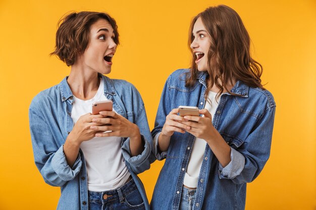 Amigos de mulheres jovens isolados sobre uma parede amarela usando telefones celulares.