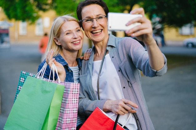 Amigos de pessoas alegres tomando selfie segurando sacos de papel com compras ao ar livre