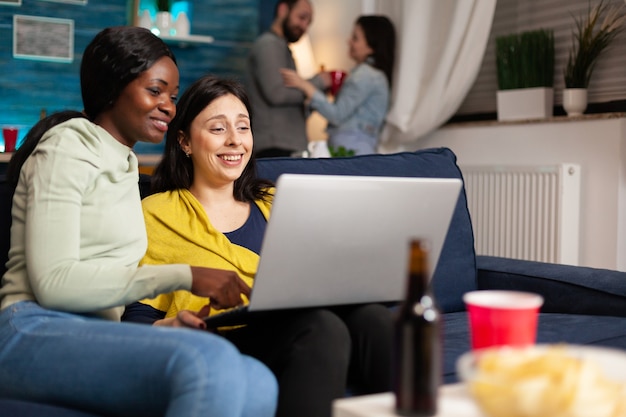 Amigos de raça mista conversando enquanto assistia a uma série de vídeos engraçados no laptop, sentado no sofá da sala. no fundo, mulher e homem bebendo cerveja, aproveitando o tempo juntos durante a festa divertida.