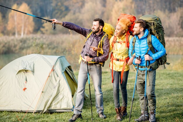 Amigos de várias etnias em jaquetas coloridas caminhando com mochilas, parados perto do acampamento no gramado verde