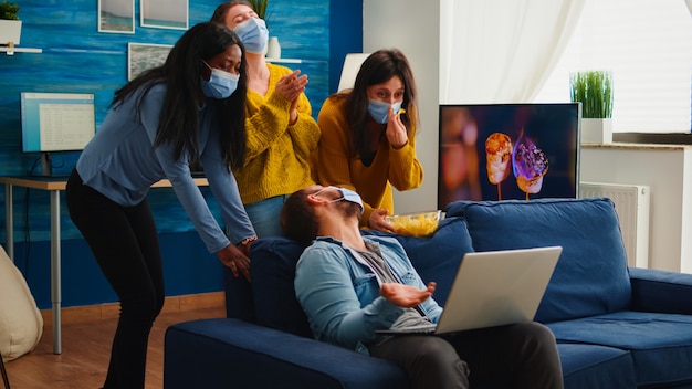 Amigos de várias etnias usando máscara facial comendo lanches usando laptop, mantendo o distanciamento social para evitar que o coronavírus se espalhe durante a pandemia global se divertindo na sala de casa. Imagem conceitual.