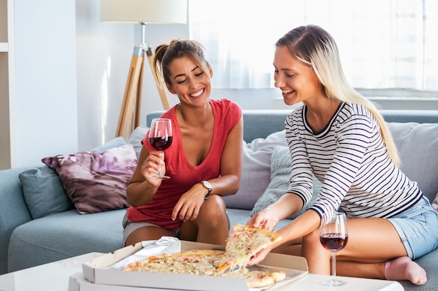 Amigos do sexo feminino comendo pizza com vinho no sofá em casa