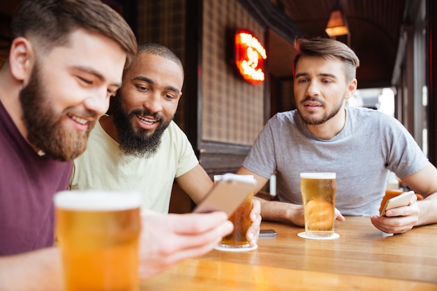 Amigos do sexo masculino felizes sentados à mesa com cerveja e usando smartphone em restaurante