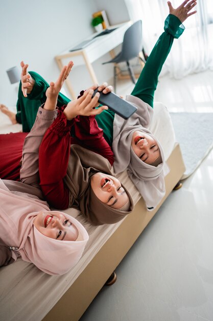 Amigos e mulheres asiáticas hijab deitam e levantam as mãos na cama enquanto tiram uma selfie juntos