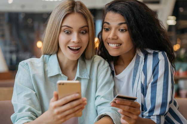 Amigos emocionais felizes segurando um cartão de crédito, usando um telefone celular, fazendo compras online em casa