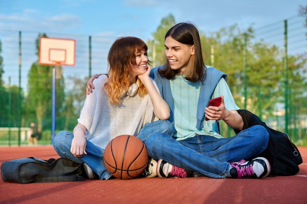 Amigos estudantes sentados na quadra de basquete ao ar livre olhando no smartphone