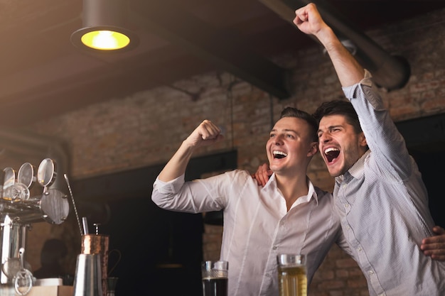 Amigos felices animando por la victoria del equipo favorito, bebiendo cerveza y celebrando el gol juntos en el pub, espacio de copia