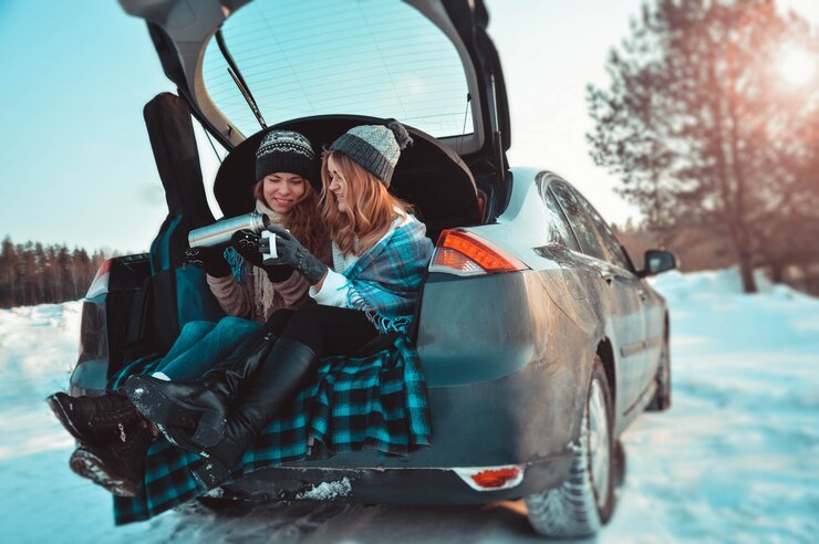 Amigos felices en el bosque de invierno en el coche. Dos niñas felices se sientan en el maletero de un coche tomando café de un termo, hablando y riendo.