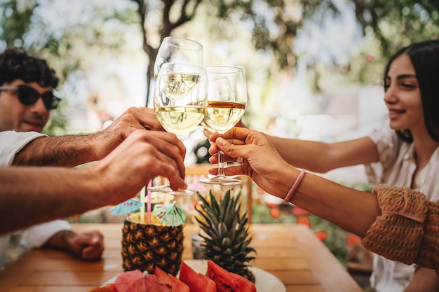 Amigos felices brindando vino blanco al aire libre - grupo de jóvenes divirtiéndose tintineando copas de vino