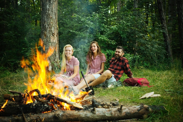 Amigos felices haciendo un picnic en el bosque. Jóvenes alegres disfrutando de las noches de verano cerca de la fogata, fin de semana en el campo